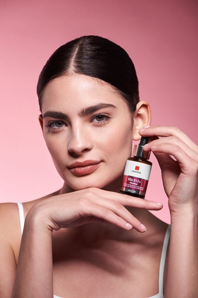A high-quality, professional product hero shot of a model holding a skin emulsion bottle against a soft pink background.